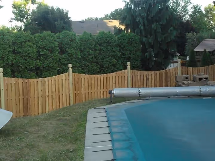 Curved wooden fence surrounding a backyard pool with greenery and trees in the background.
