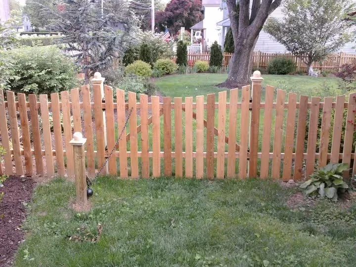 Wooden picket fence with gate enclosing a grassy backyard with trees and bushes.