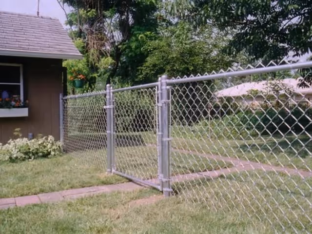 Chain-link fence gate surrounding a yard with grass and a house corner in the background.