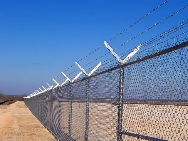 Chain-link fence topped with angled barbed wire under a clear blue sky over dry ground.