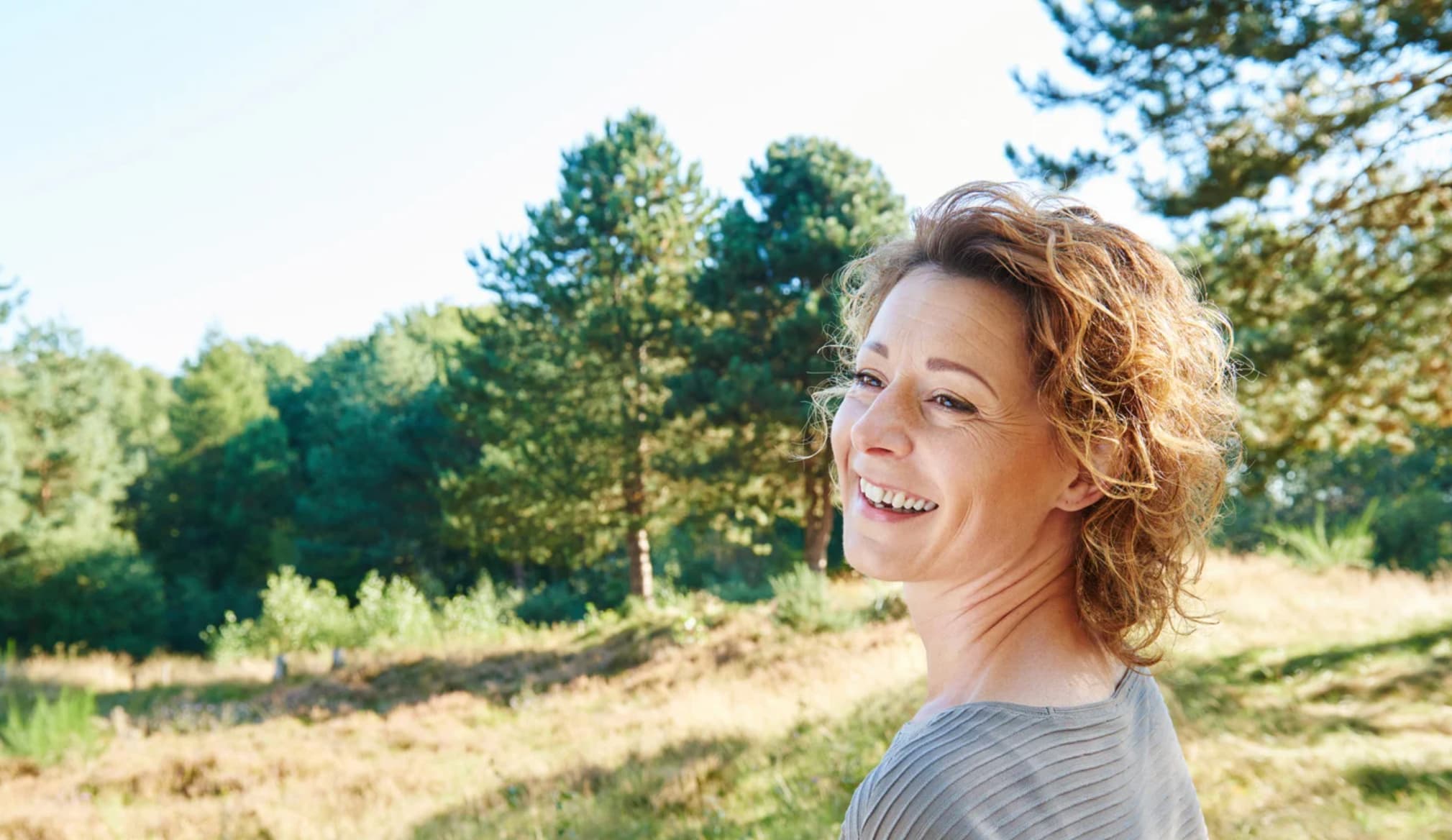 Smiling woman with curly hair stands outdoors with trees and grasses in the background.