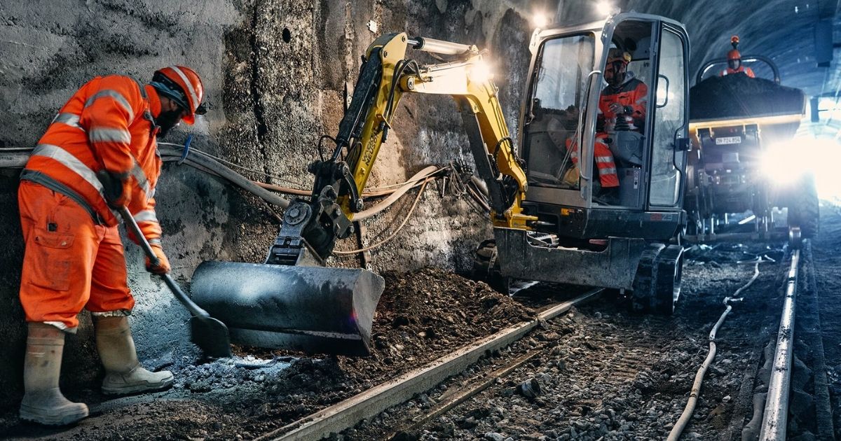 Bauarbeiter in orangefarbener Schutzkleidung arbeitet mit einem Mini-Bagger und Schaufel in einem Tunnel mit Bahnschienen.