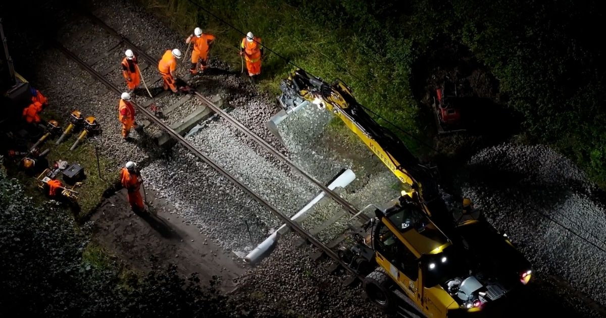 Nachtszene mit mehreren Arbeitern in orangenen Schutzanzügen, die Gleisbauarbeiten mit schwerem Gelbbaumaschinen an einer Eisenbahnstrecke durchführen.