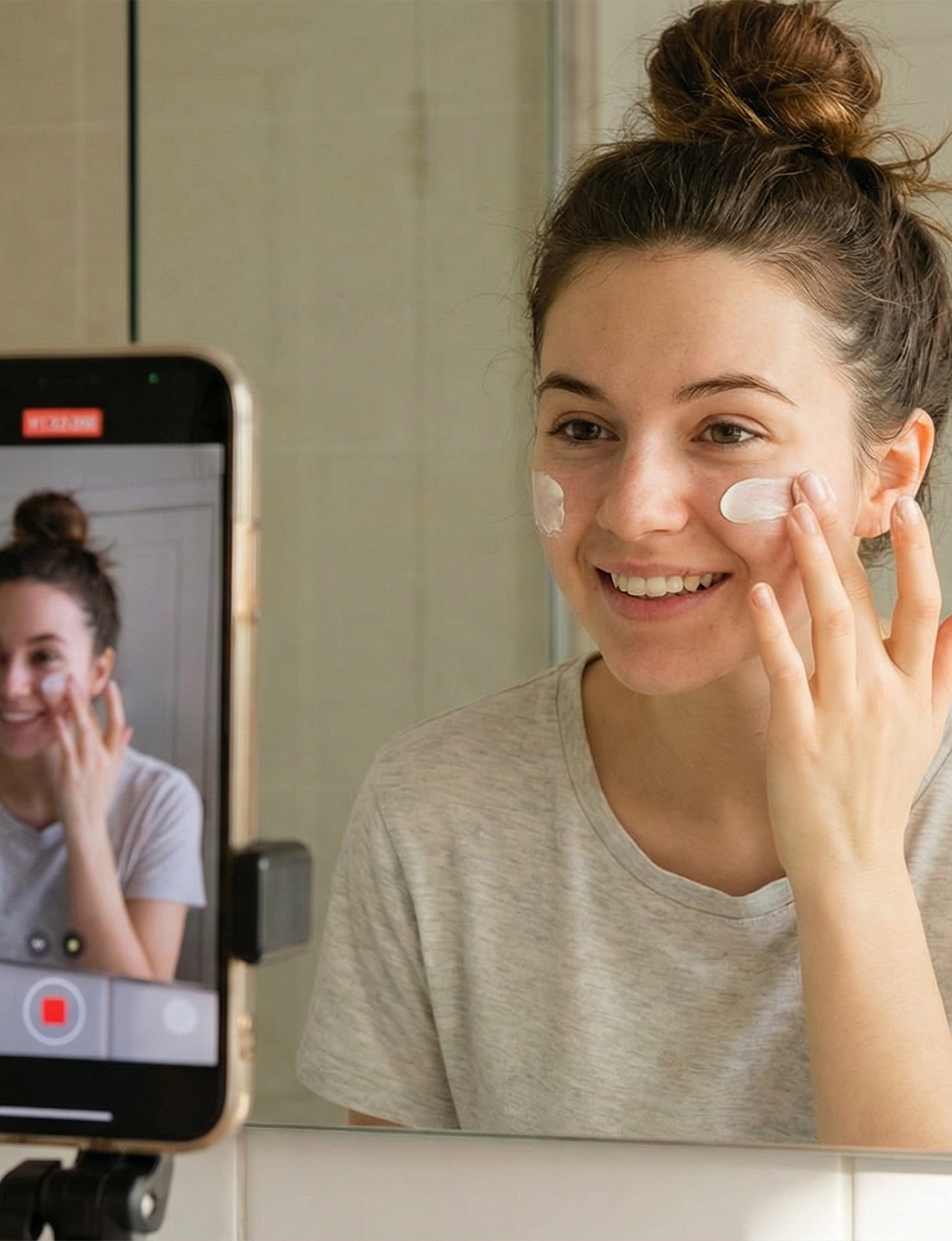Young woman with hair in a bun smiling while applying cream to her cheeks in front of a mirror, recording herself with a smartphone.