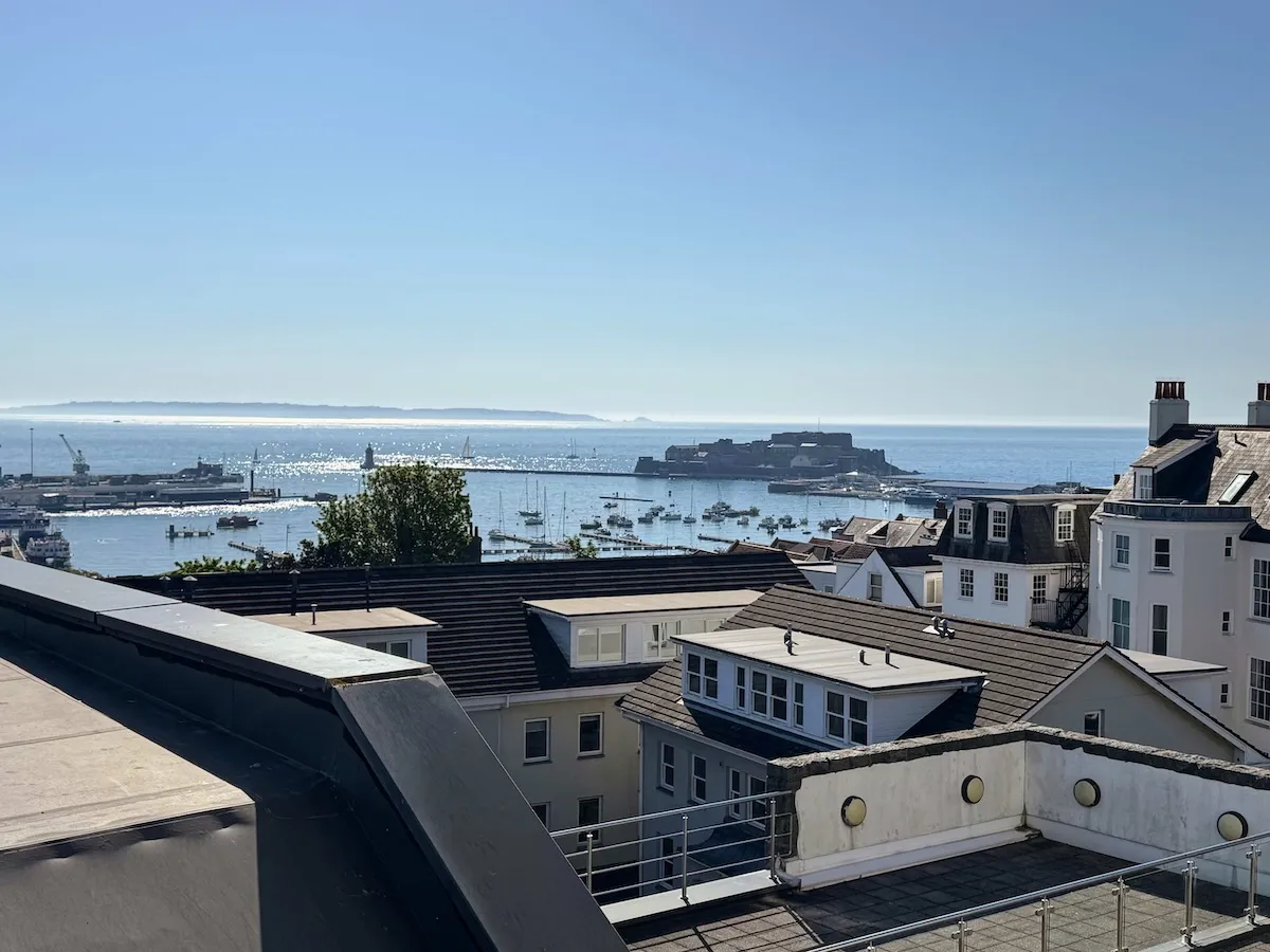 View over rooftops toward a harbor with boats and a distant island under a clear blue sky.