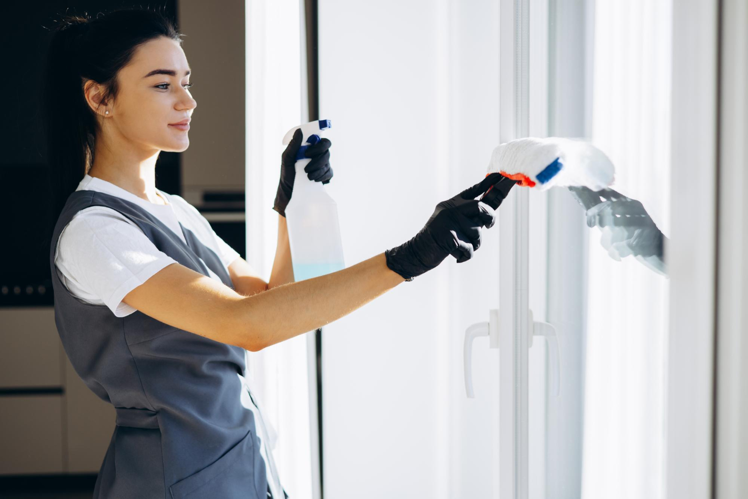 Woman wearing gloves cleaning a glass window with a spray bottle and a microfiber mop.