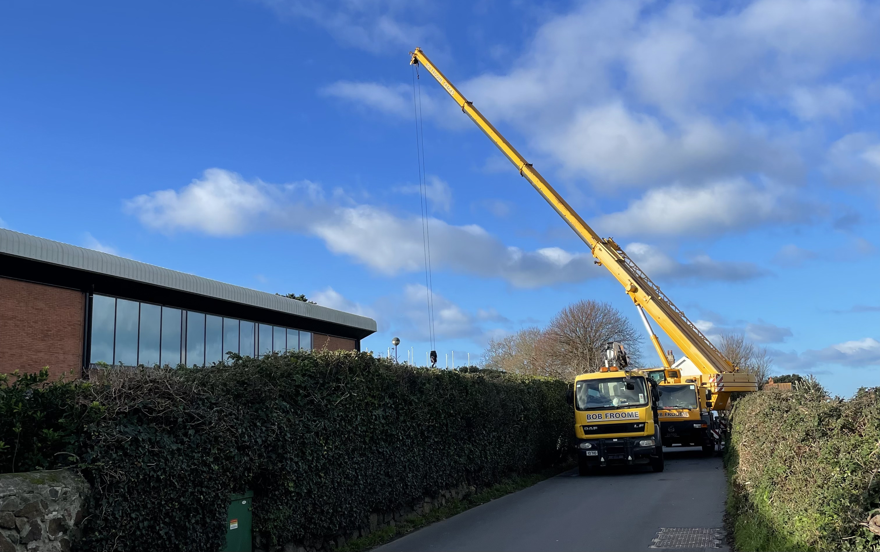 Yellow construction crane and truck labeled 'BOB FROOME' on a narrow road next to a hedge and a brick building with large windows.