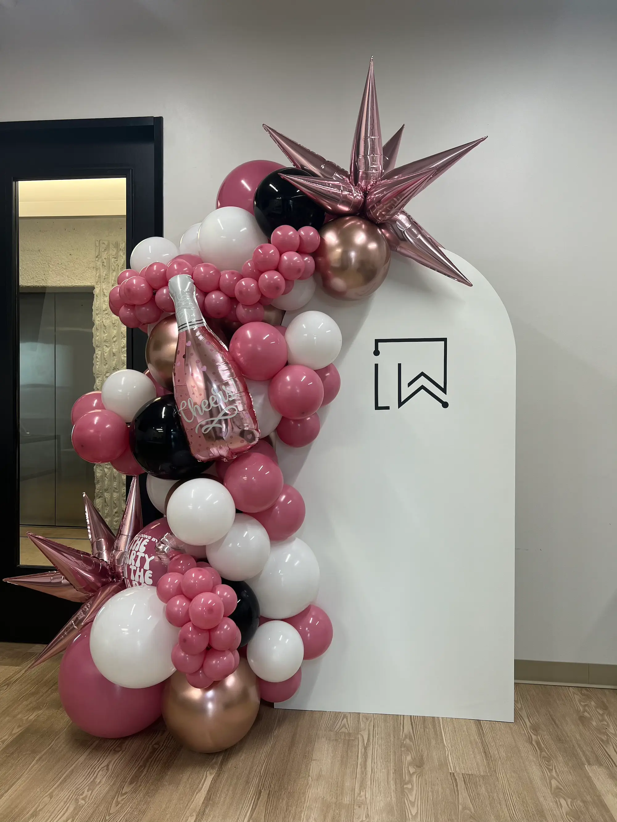 Decorative balloon arrangement in pink, white, black, and rose gold colors with starburst balloons and a balloon shaped like a champagne bottle beside a white arch backdrop for dentist office