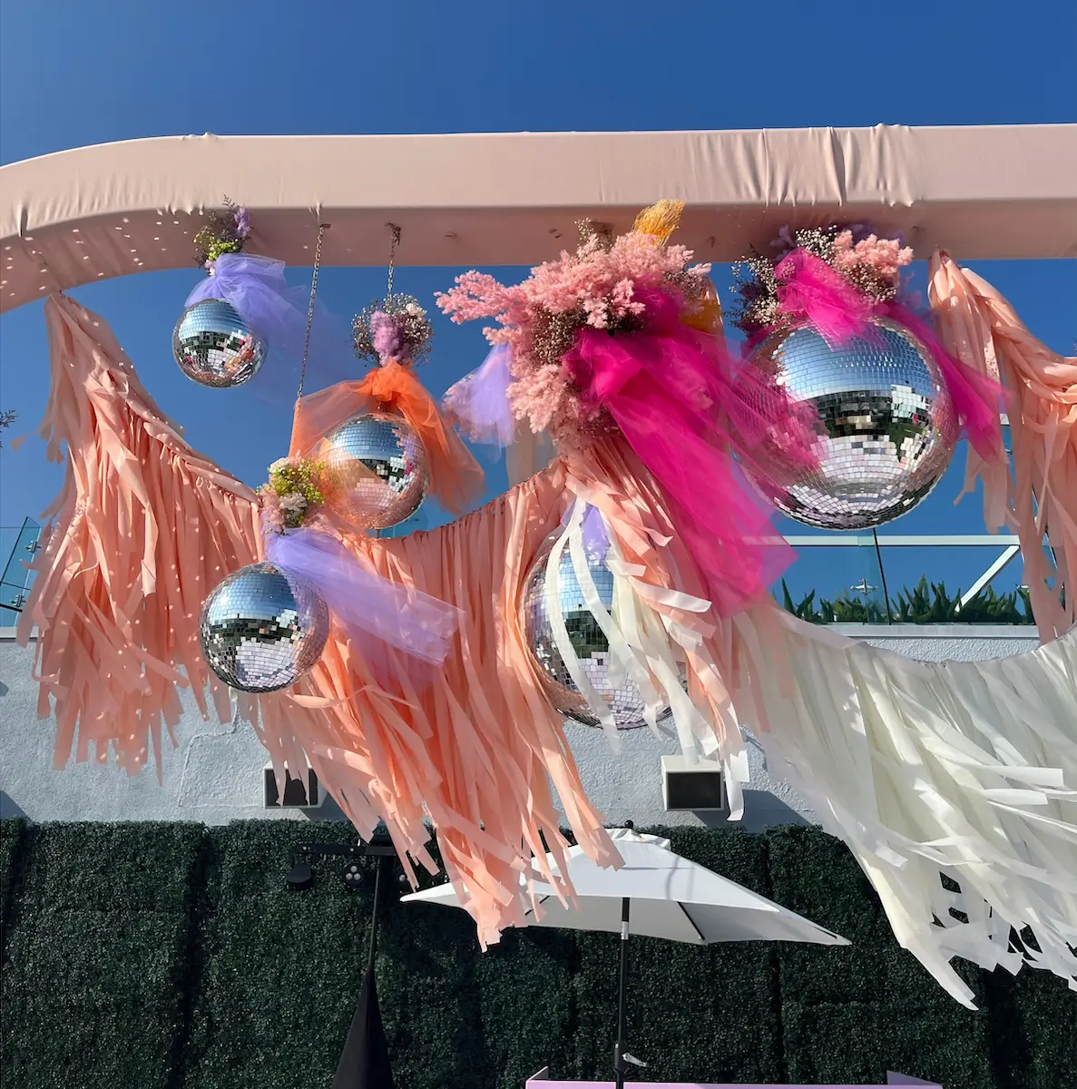 Wedding Outdoor decoration with hanging disco balls adorned with colorful tulle and flowers, peach and white fringe garlands, under a clear blue sky in Dallas, TX.