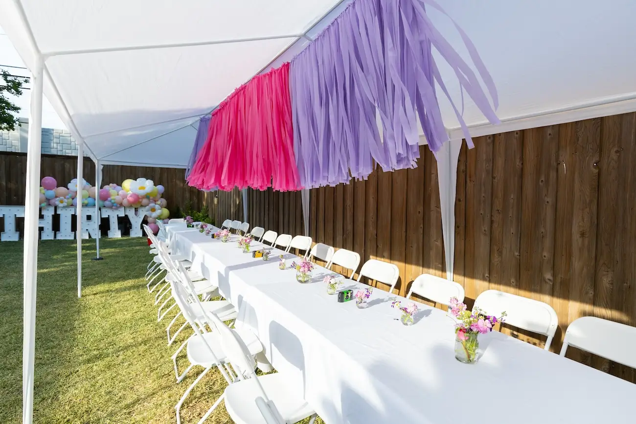 Long outdoor table with white tablecloth and white chairs under a tent, decorated with small flower vases and pink and purple hanging fringe in Dallas.