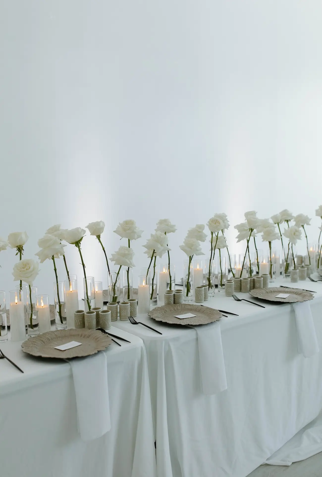 Minimalist table setup with white roses in glass vases, white candles, beige plates, and black utensils on a white tablecloth.
