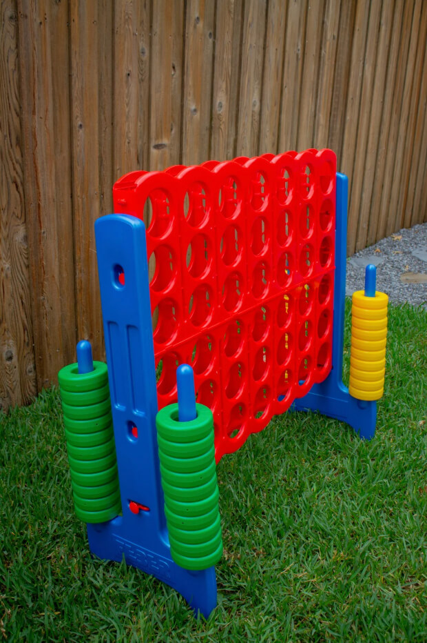 Outdoor oversized Connect 4 game rental with red grid, blue frame, green and yellow discs on grass beside a wooden fence in Dallas