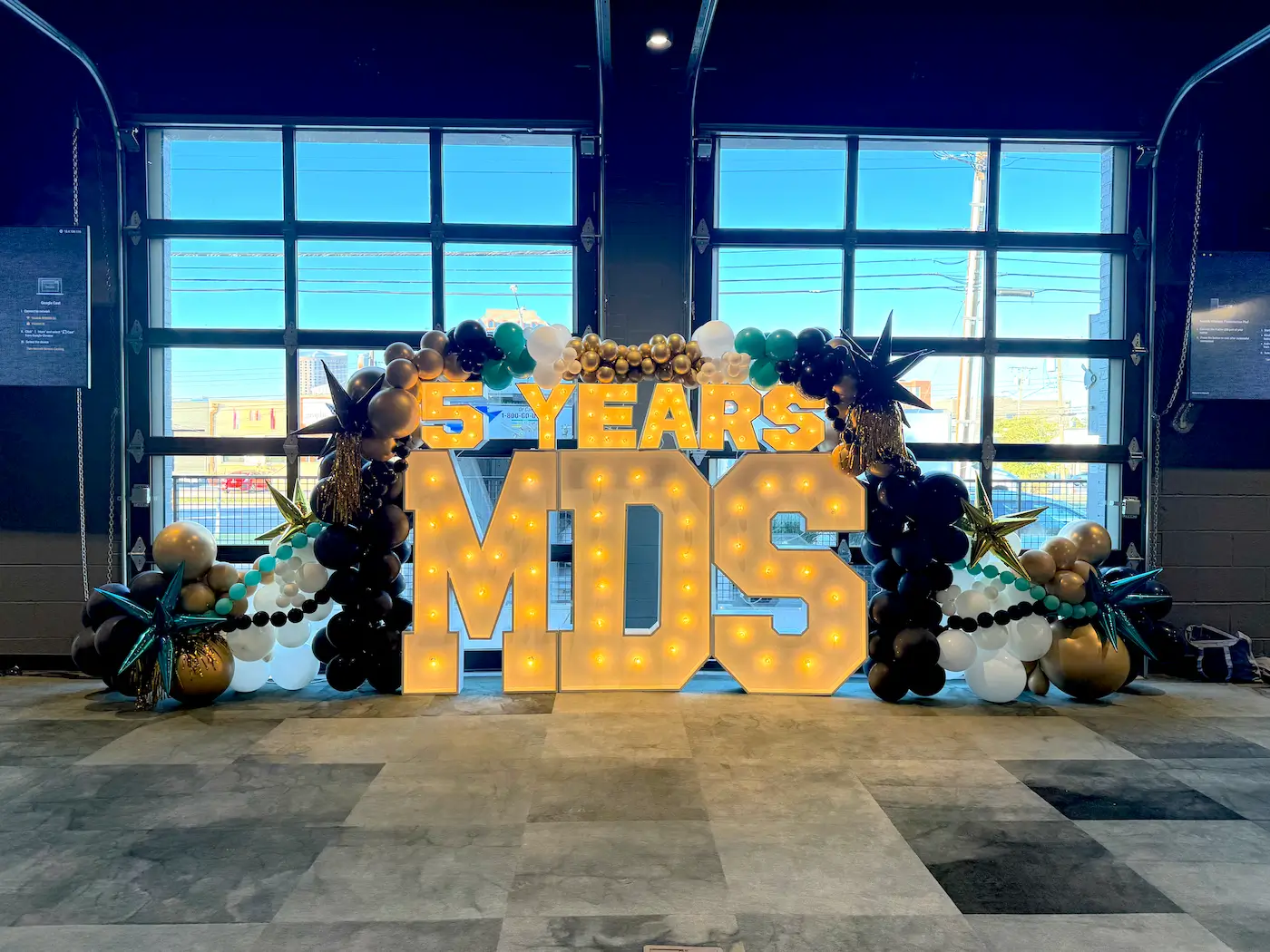 Illuminated marquee letters spelling 5 YEARS MDS decorated with black, gold, white, and teal balloons inside a building with large garage-style windows.