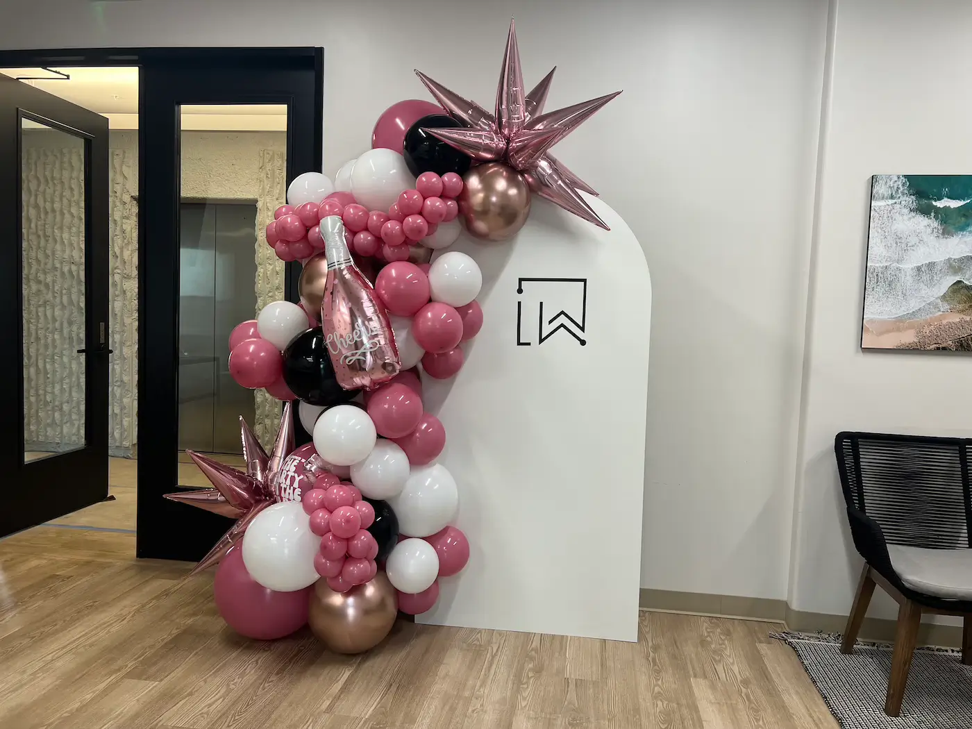 Decorative balloon arch in pink, white, black, and metallic colors with a champagne bottle balloon saying 'cheers' next to a white panel with a minimalist logo, inside an office in Lakewood neighborhood in Dallas.