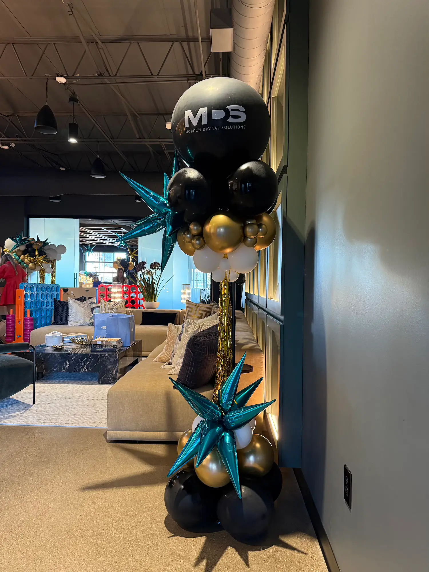 Black, gold, white, and teal balloon arrangement with a large black balloon displaying the text 'MDS Moroch Digital Solutions' in a modern lounge area in Dallas Design District.