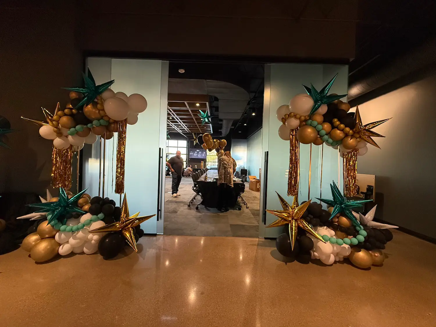 Balloon arch decorations in gold, black, white, teal, and metallic star shapes framing double glass doors leading to a conference room with people inside at Dallas' Design District.