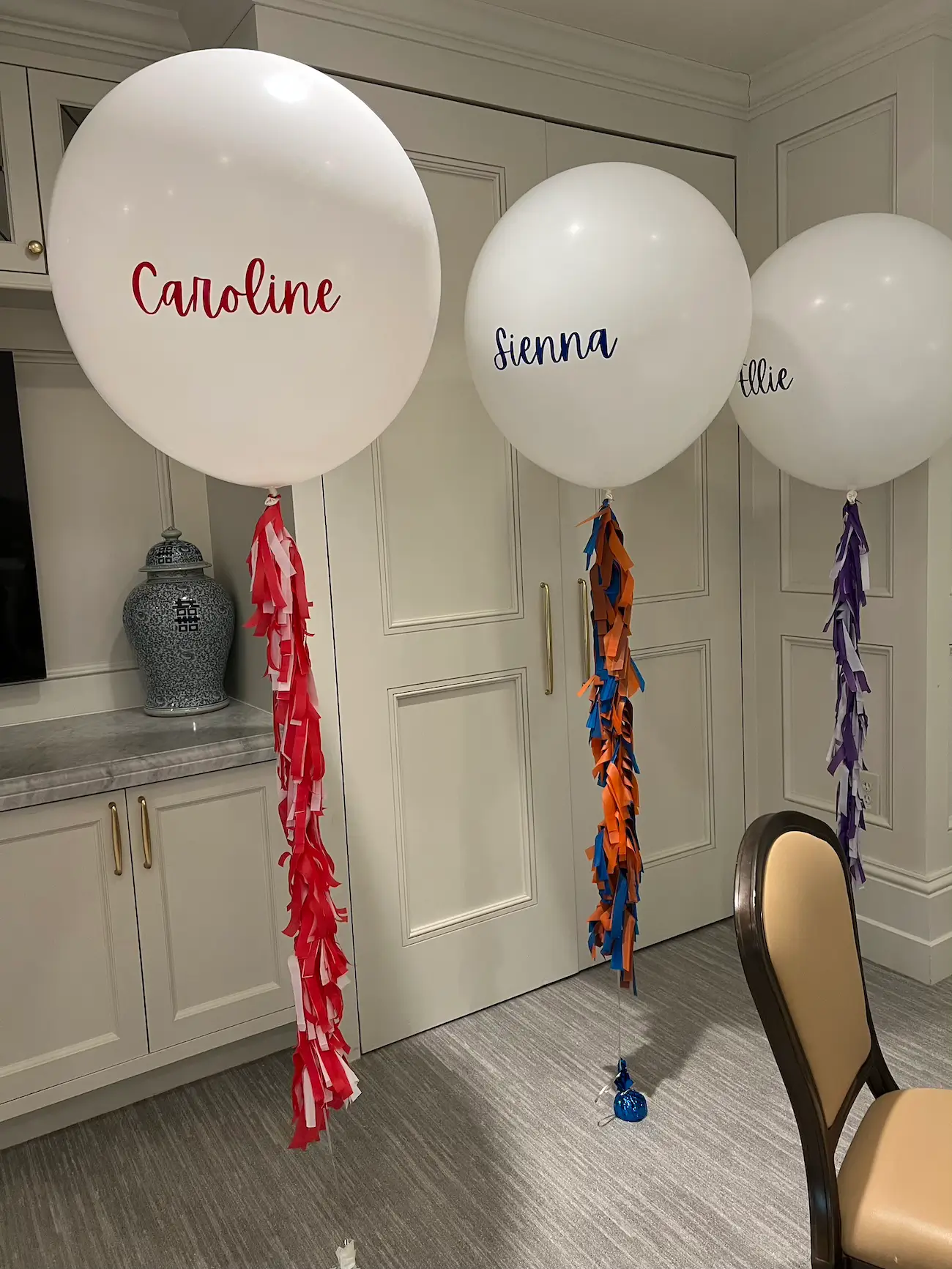 Three large white helium balloons with names Caroline, Sienna, and Ellie, each tied with colorful tassel tails in red, orange and blue, and purple, inside a room with beige walls at Dallas Country Club.