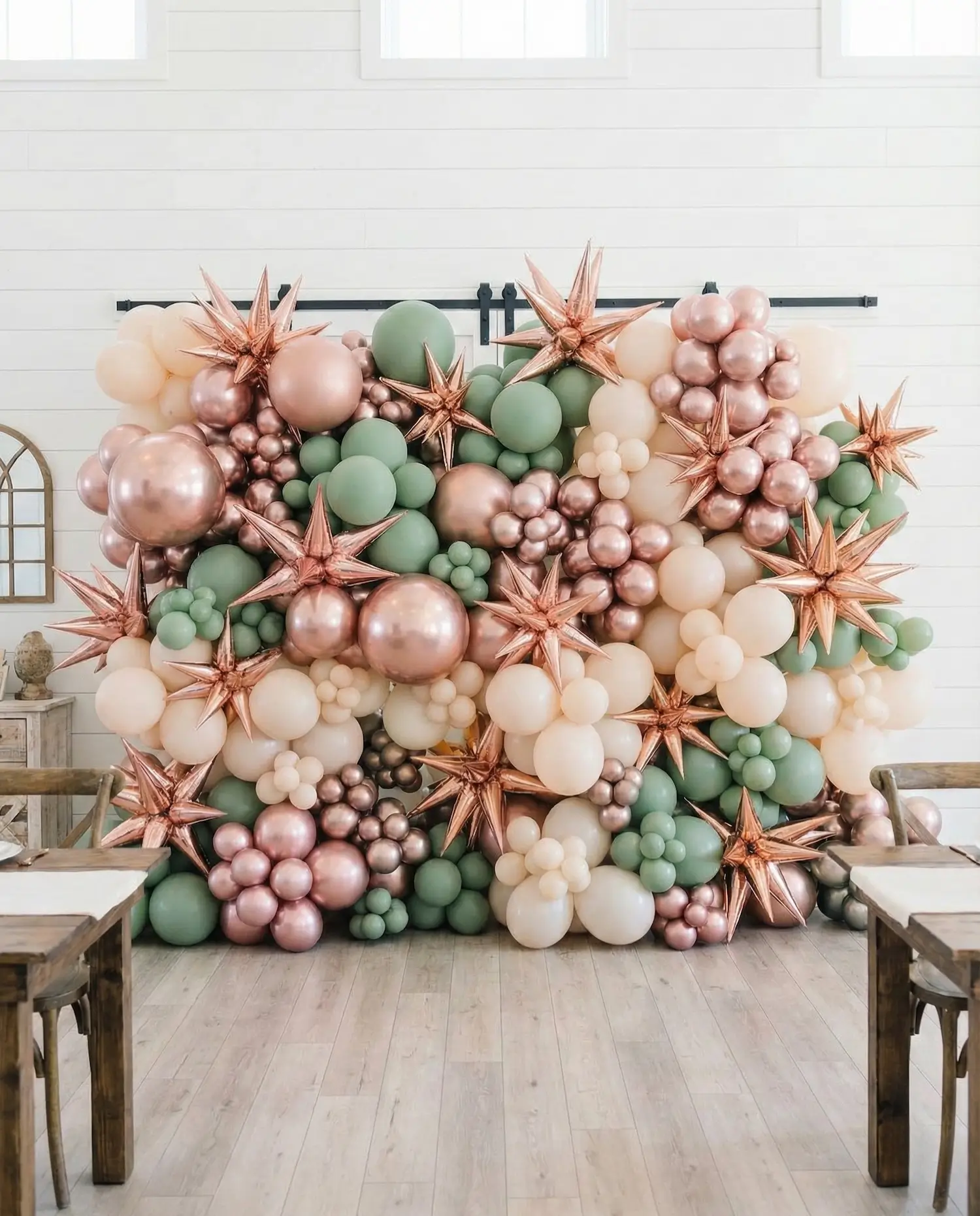 Decorative balloon wall made of clusters of cream, green, and rose gold balloons with rose gold star-shaped balloons in a room with wooden tables and chairs in Dallas.