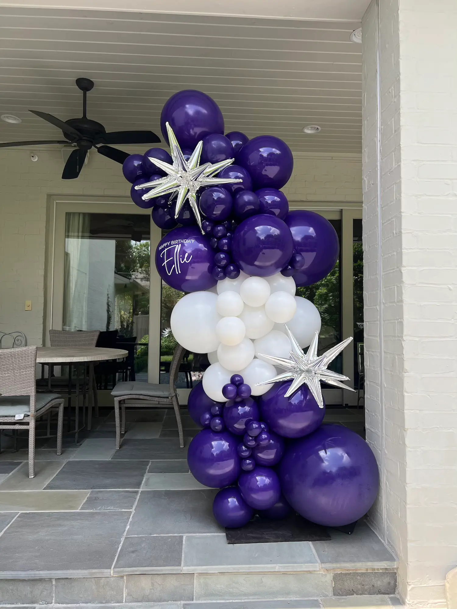 Purple and white balloon column with silver star-shaped balloons and a purple balloon reading Happy Birthday Ellie on a patio in University Park Dallas.