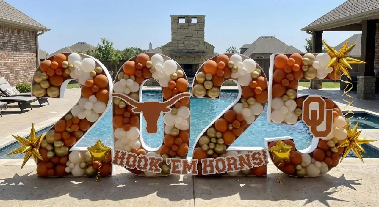 Outdoor backdrop with repeated text 'texas aggie' and one highlighted 'texas aggies' in maroon, decorated with maroon, beige, and pink balloon clusters on the right side in Dallas.