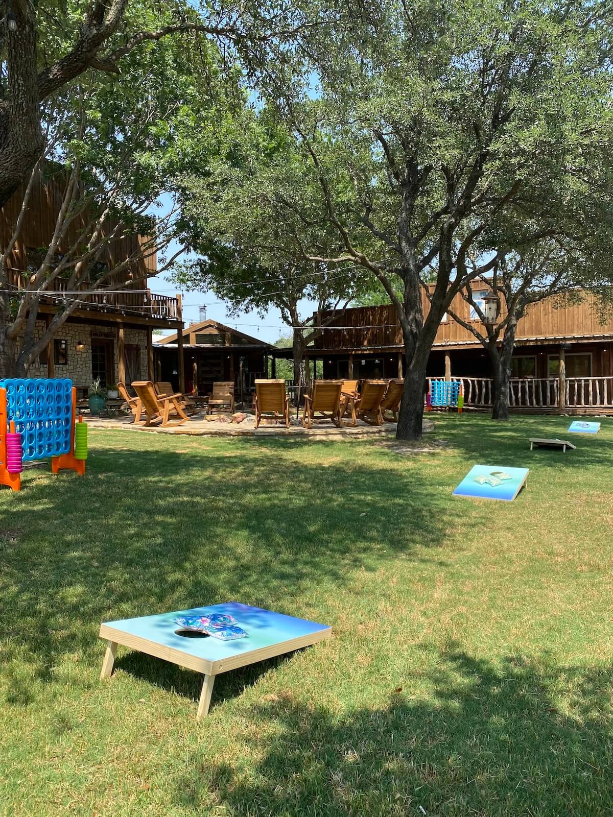Outdoor lawn with cornhole boards set up on grass, surrounded by trees and wooden chairs near rustic buildings.