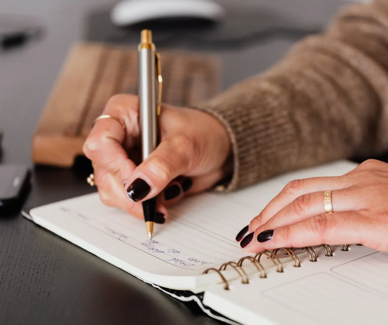 Person with dark nail polish writing in a spiral-bound notebook with a pen beside a computer keyboard.