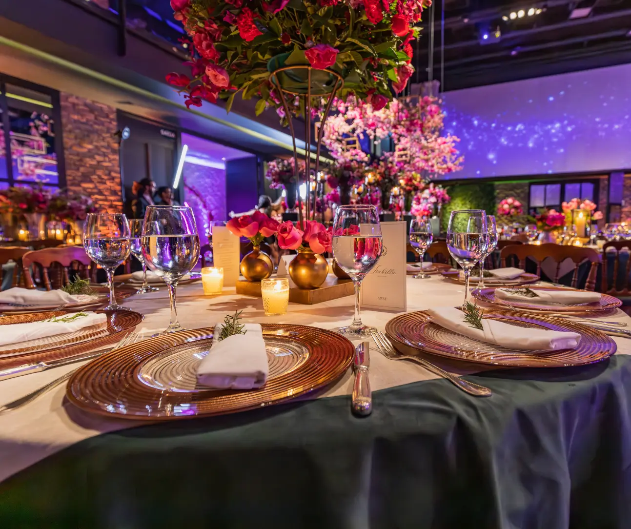 Elegant table setting with glass plates, white napkins, and floral centerpieces with red and pink roses in a warmly lit event space.
