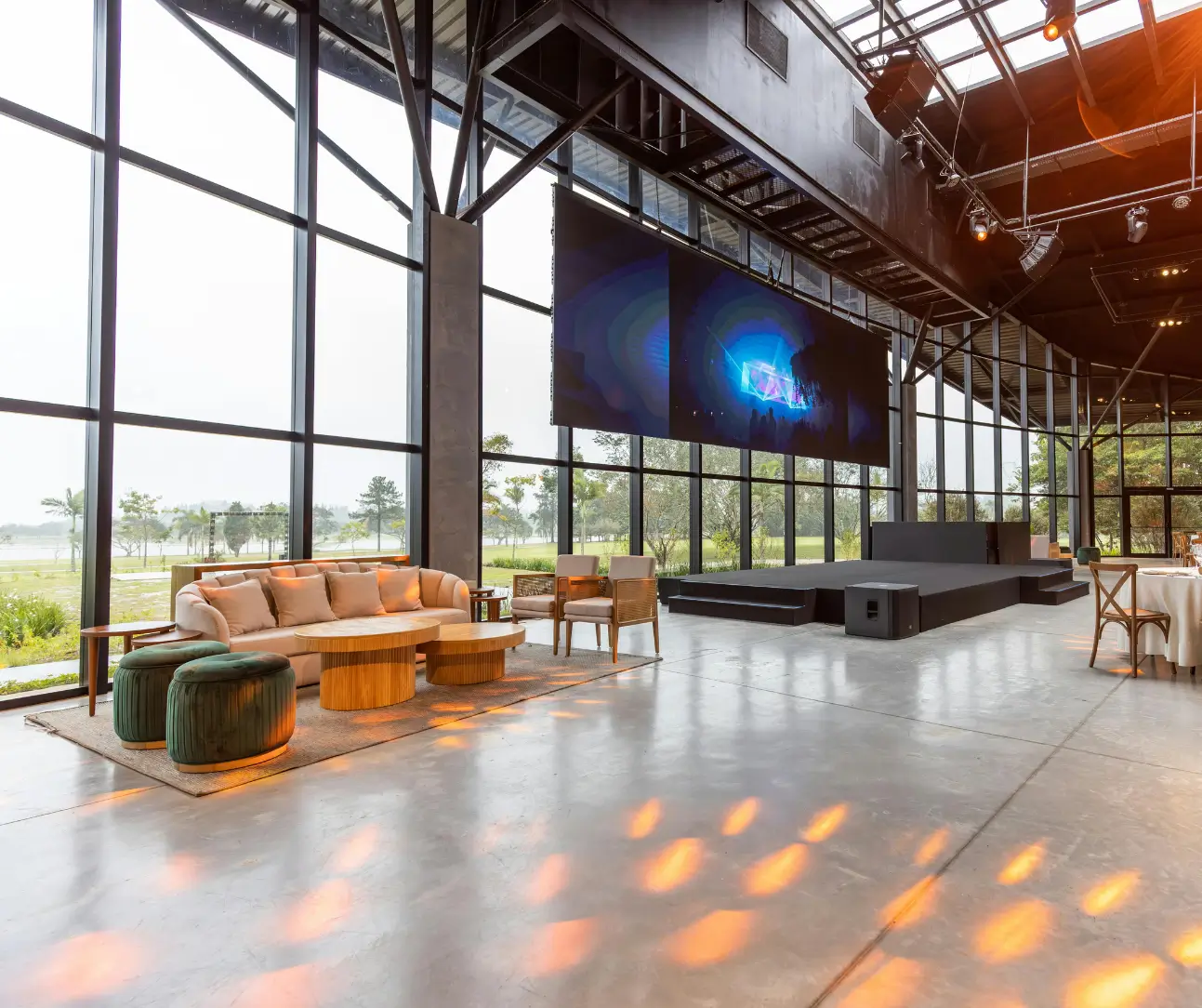 Spacious event venue with large floor-to-ceiling windows, modern seating area, raised stage, and large hanging screen.