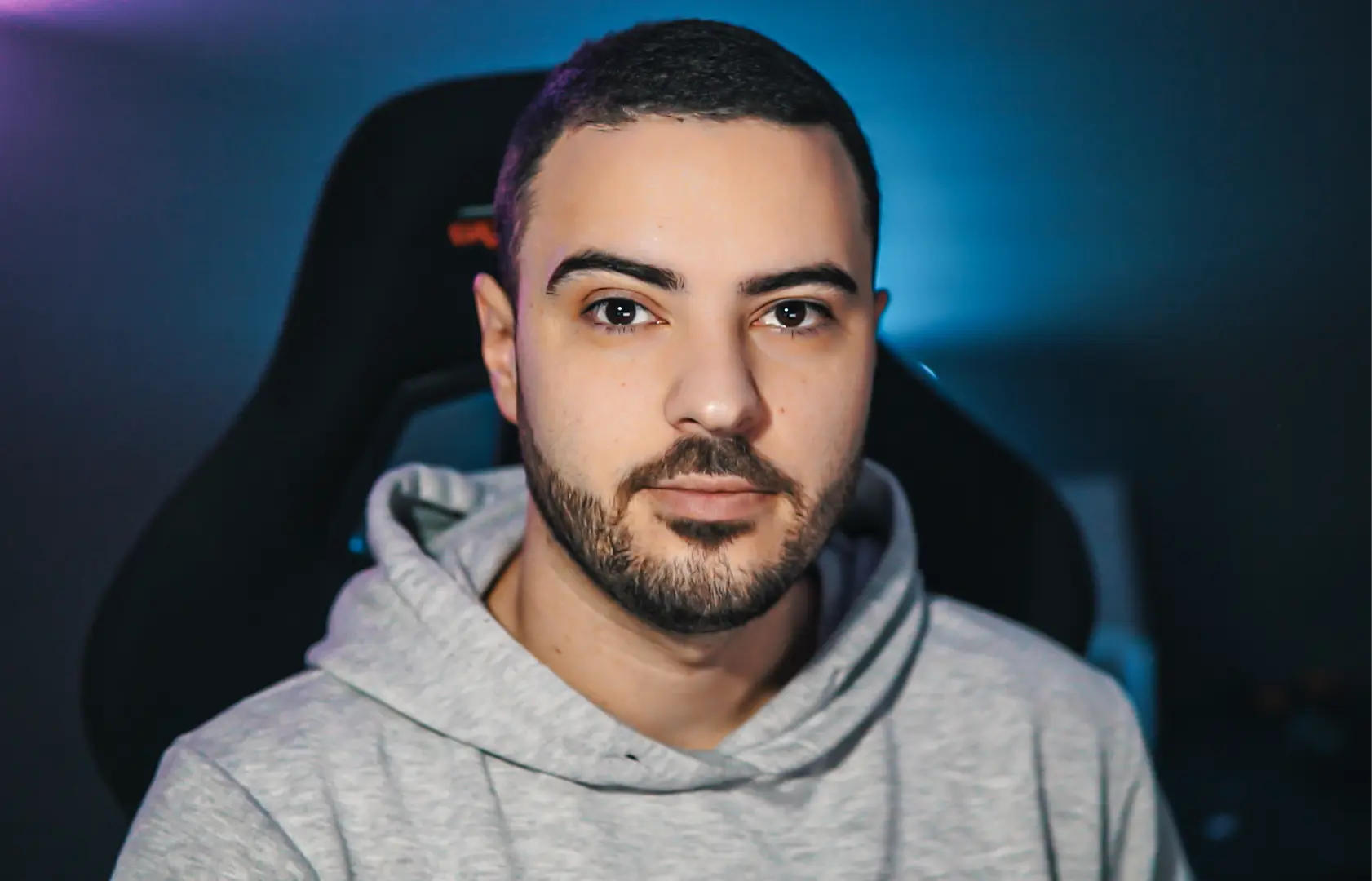 Young man with short dark hair and beard wearing a gray hoodie seated in a black gaming chair with blue background lighting.