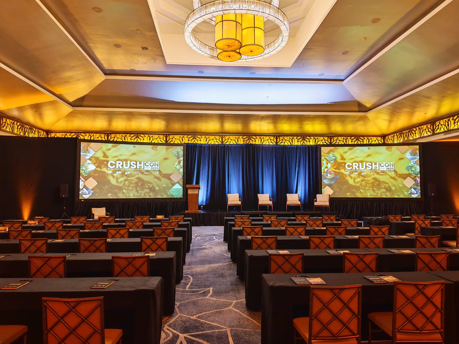 Conference room set up with rows of tables and chairs facing a stage with four chairs, a podium, and two large screens displaying 'CRUSHCON 2025'.