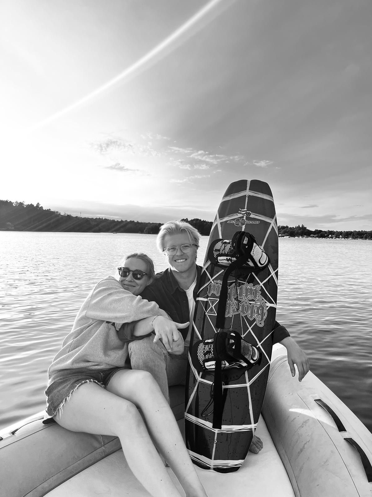 Smiling young couple sitting on a boat by the water at sunset, holding a wakeboard.