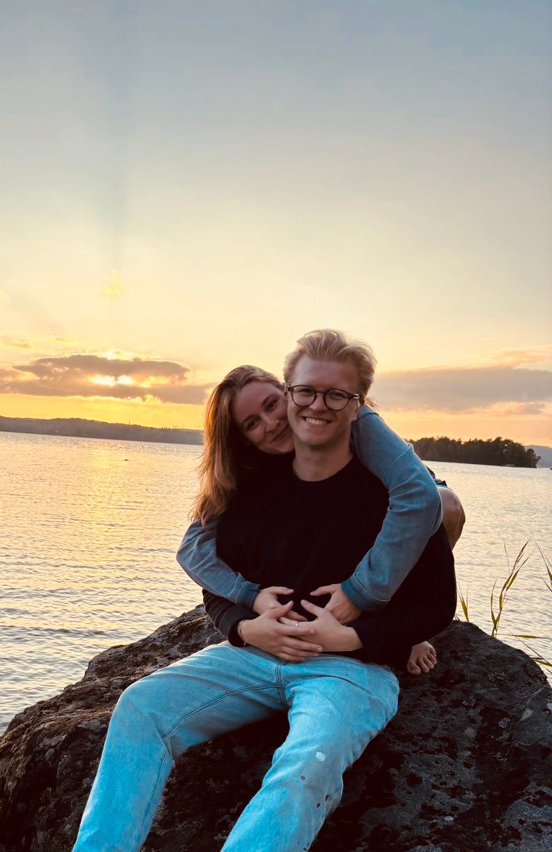 Young couple sitting on a rock by a lake at sunset, the woman hugging the man from behind and both smiling.