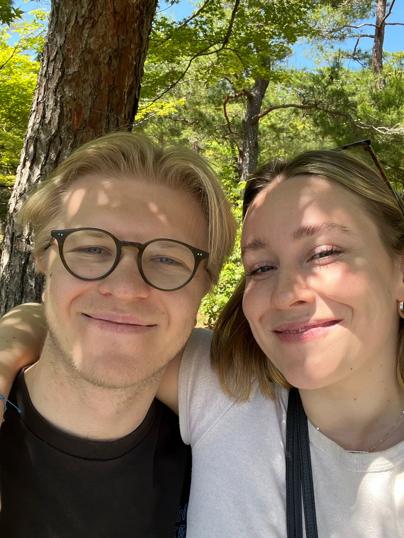 Smiling couple taking a close-up selfie outdoors with trees and sunlight casting shadows on their faces.