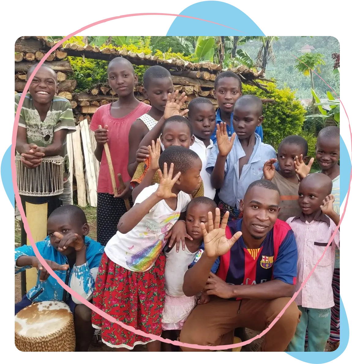 A group of children and a man outdoors waving and smiling, with stacked firewood and lush greenery in the background.
