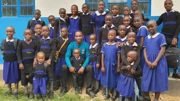 Group of smiling schoolchildren and a teacher standing outside a building, wearing blue uniforms.