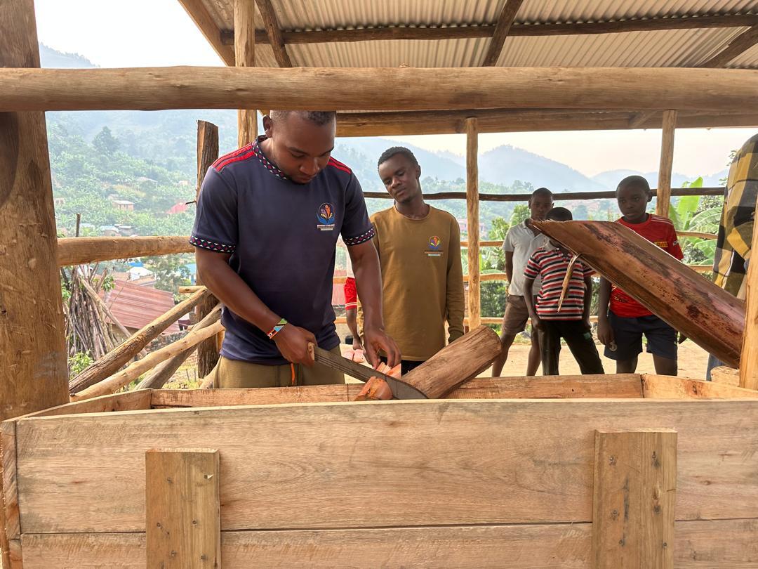 A young man saws a piece of wood while others watch inside a wooden structure against a mountainous backdrop.