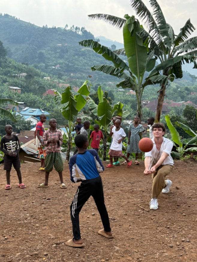 A group of children and a young man playing volleyball outdoors on a dirt ground with green hills and banana trees in the background.