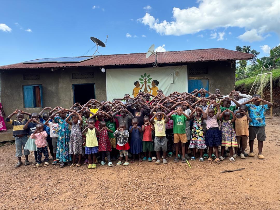 Large group of children and adults standing outside a building, forming heart shapes with their hands under a blue sky.