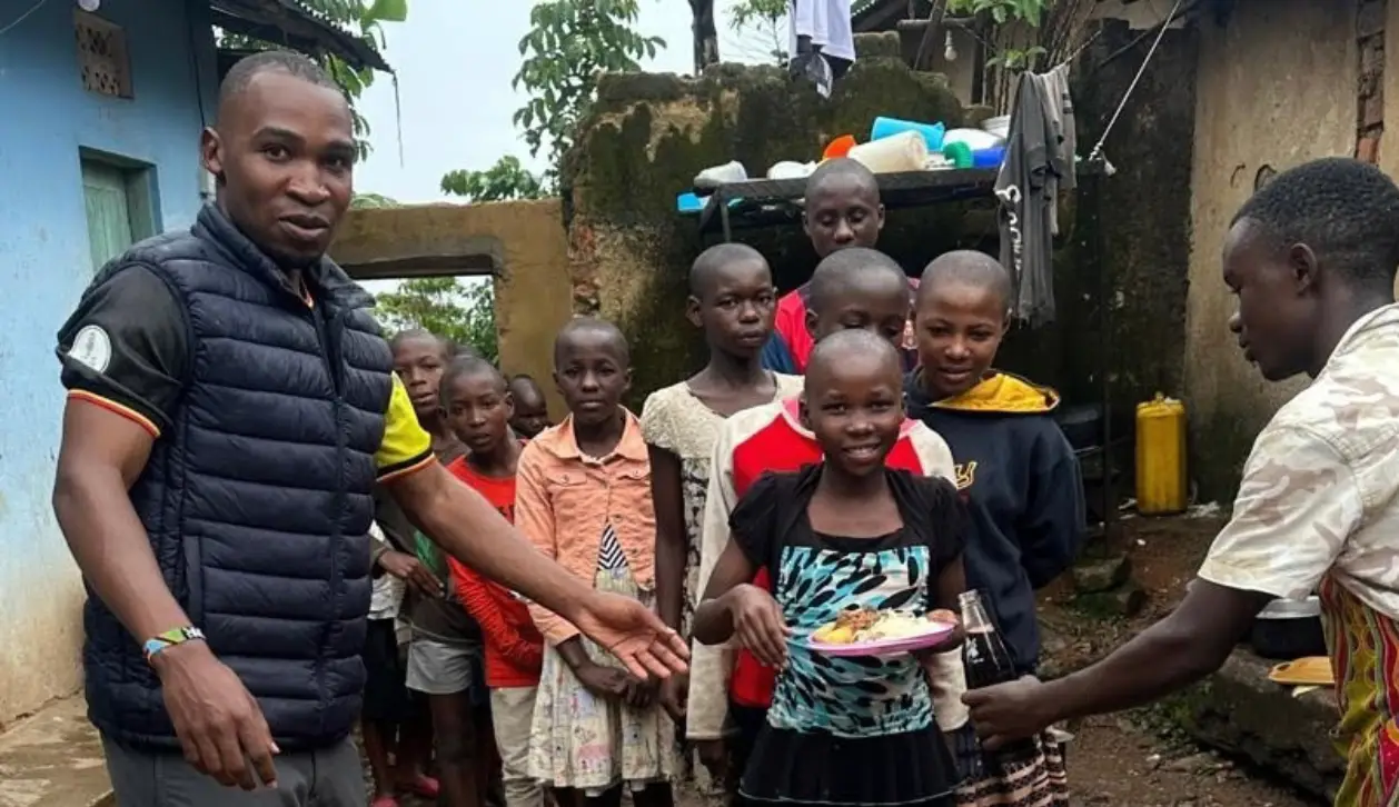 A group of children standing outside with one girl smiling and holding a plate of food, while an adult hands a bottle to another child.
