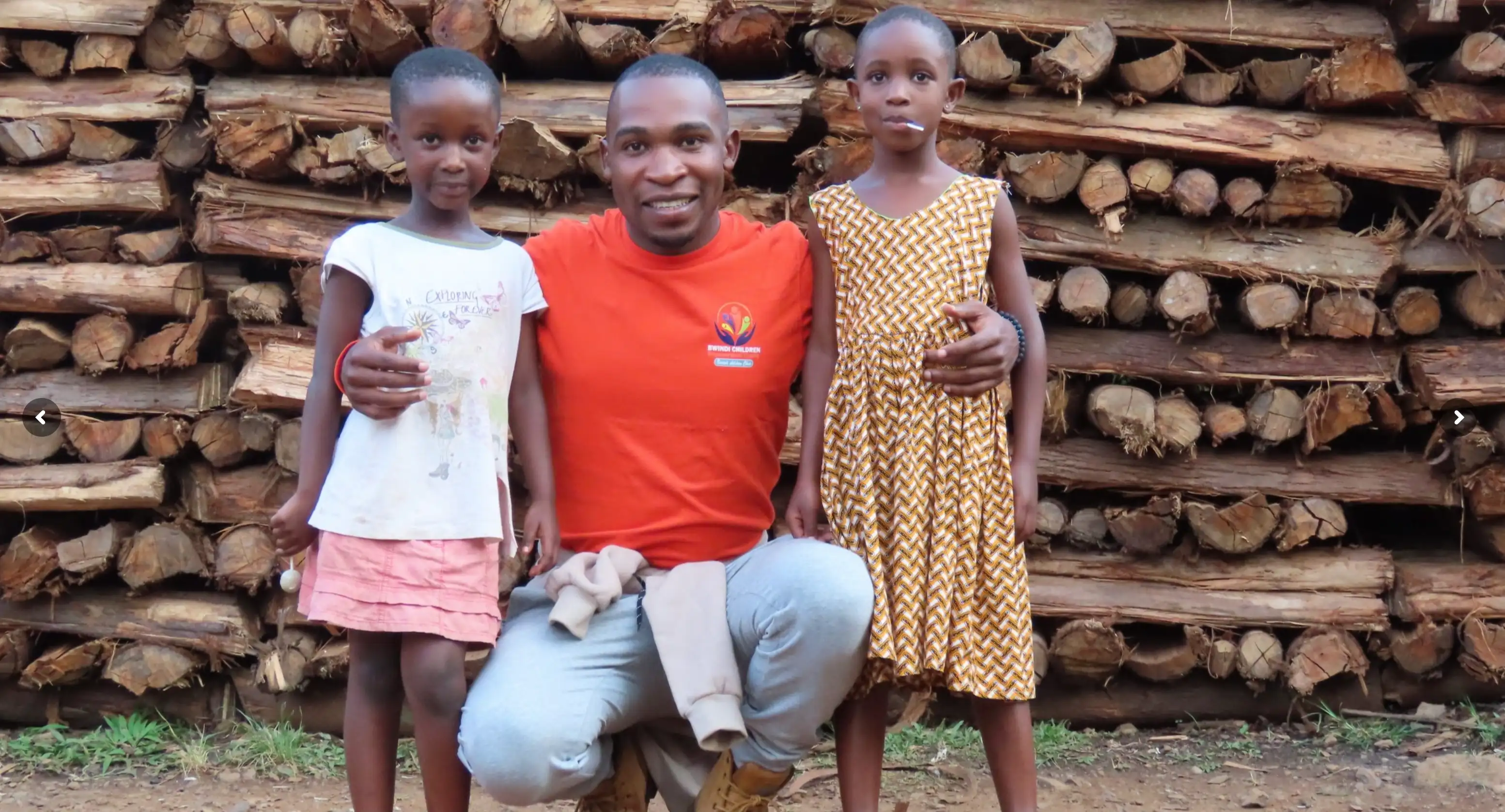 Man in orange shirt crouching with arms around two young girls standing in front of stacked firewood.