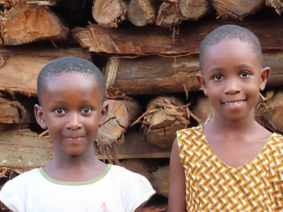 Two smiling young African girls standing in front of stacked logs, one wearing a white shirt and the other a patterned yellow dress.