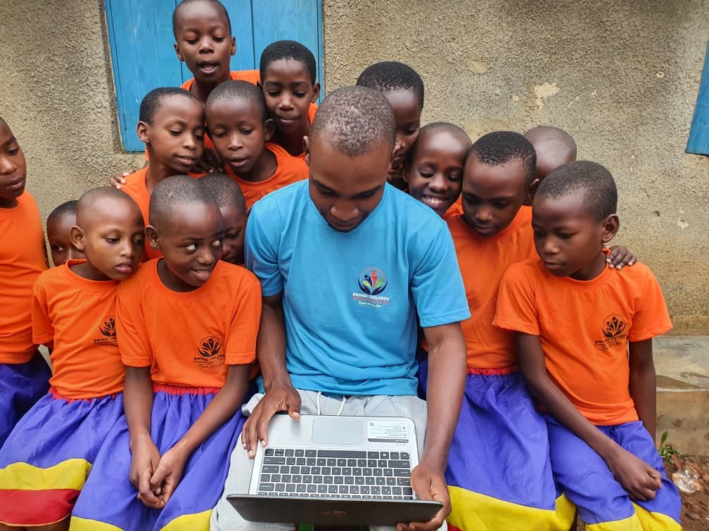 Group of children in orange shirts and purple skirts gathered closely around a man in a blue shirt using a laptop outdoors.