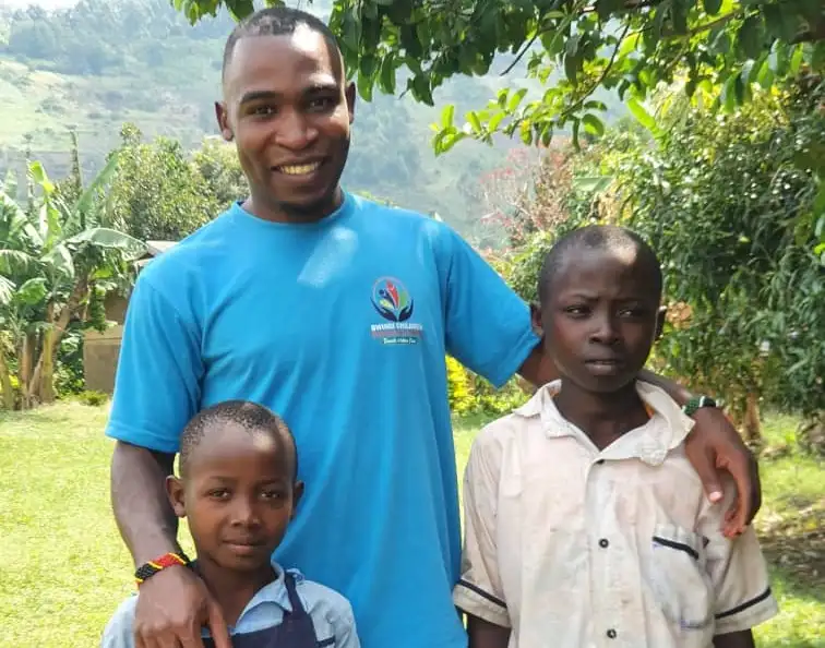 Smiling man in a blue shirt with an arm around two boys standing outdoors with greenery and hills in the background.