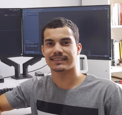 Homem jovem sorrindo em frente a dois monitores de computador em um ambiente de escritório com estantes de livros ao fundo.
