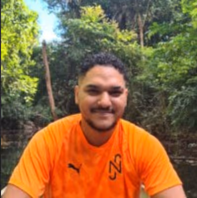 Homem sorridente de camiseta laranja com logo e fundo de vegetação.