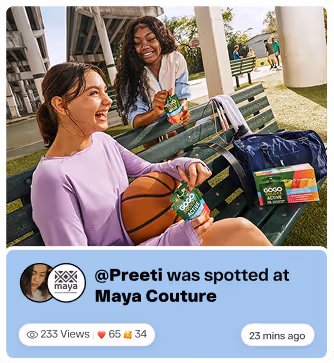 Two women sitting on a green bench outdoors, smiling and holding GoGo squeeZ applesauce pouches, with a basketball and personal items beside them.