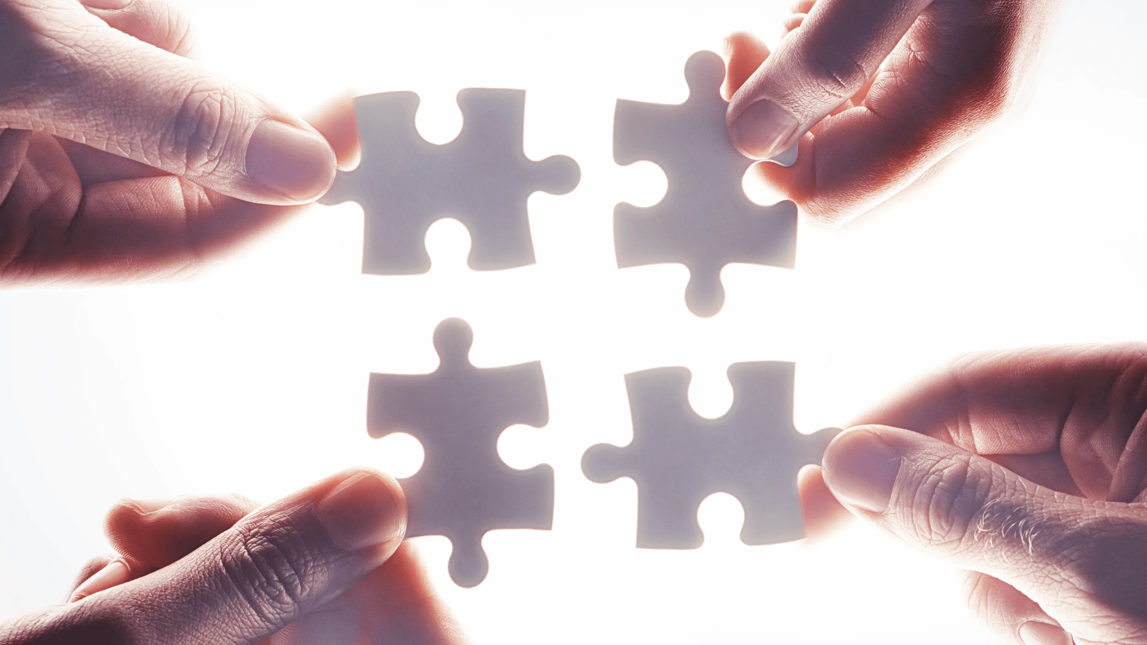 Four hands each holding a puzzle piece coming together to form a square.