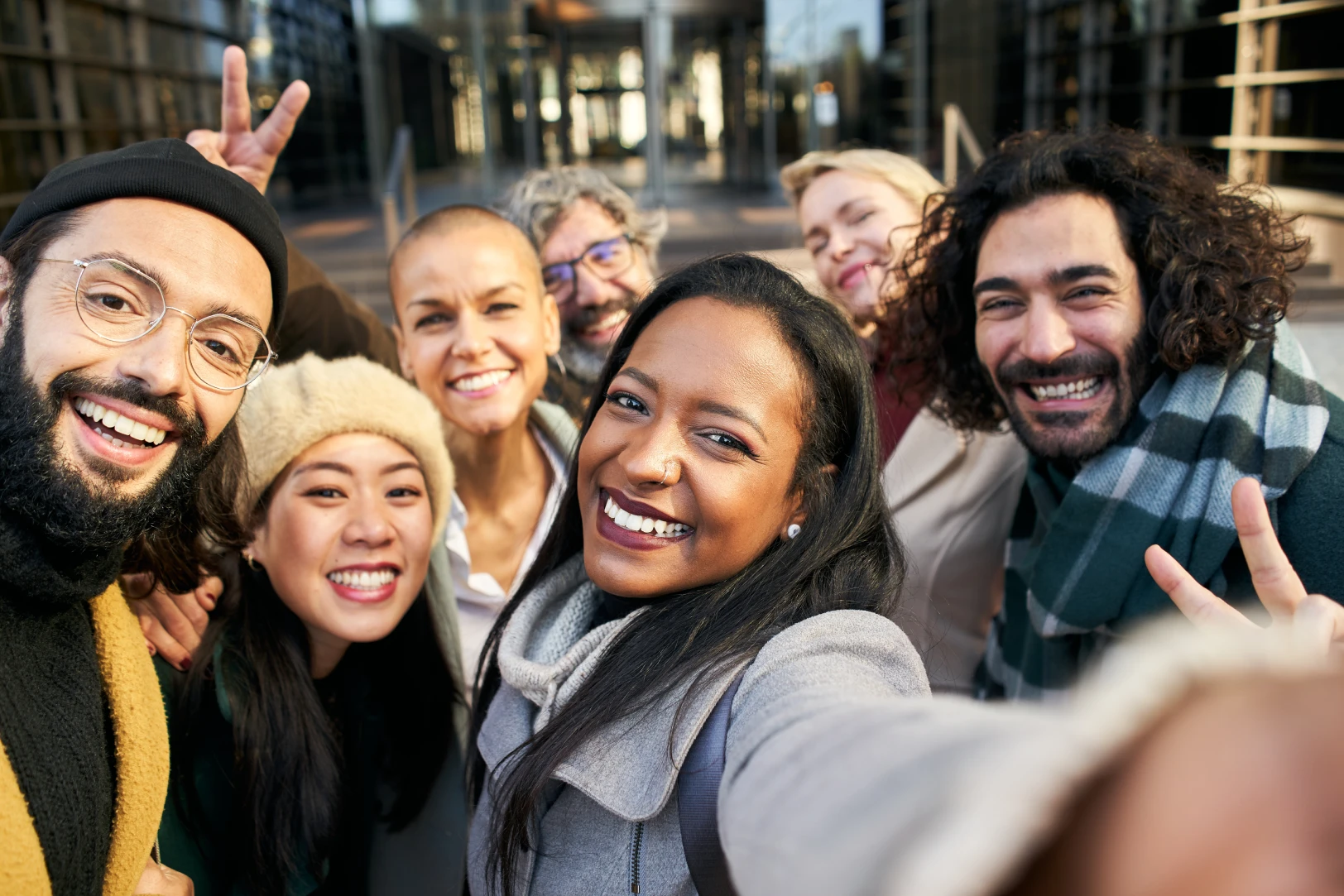A diverse group of seven friends smiling and posing for a selfie outdoors in front of a building.