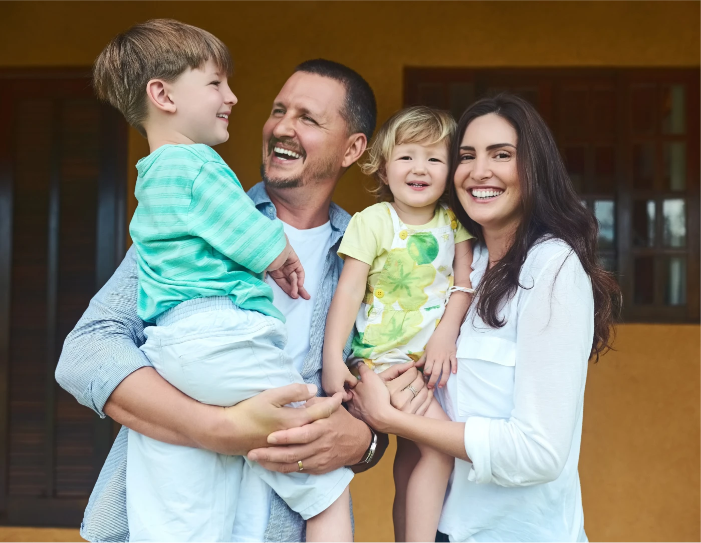 Smiling family of four with father holding a boy and mother holding a toddler, standing in front of a house.