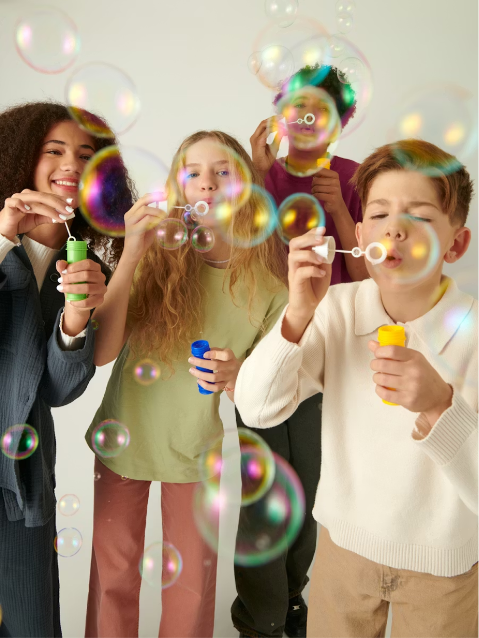 Four diverse teenagers blowing colorful soap bubbles indoors against a light background.